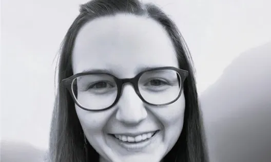 A black and white portrait of a smiling person wearing glasses, with shoulder-length hair, set against a blurred outdoor background.
